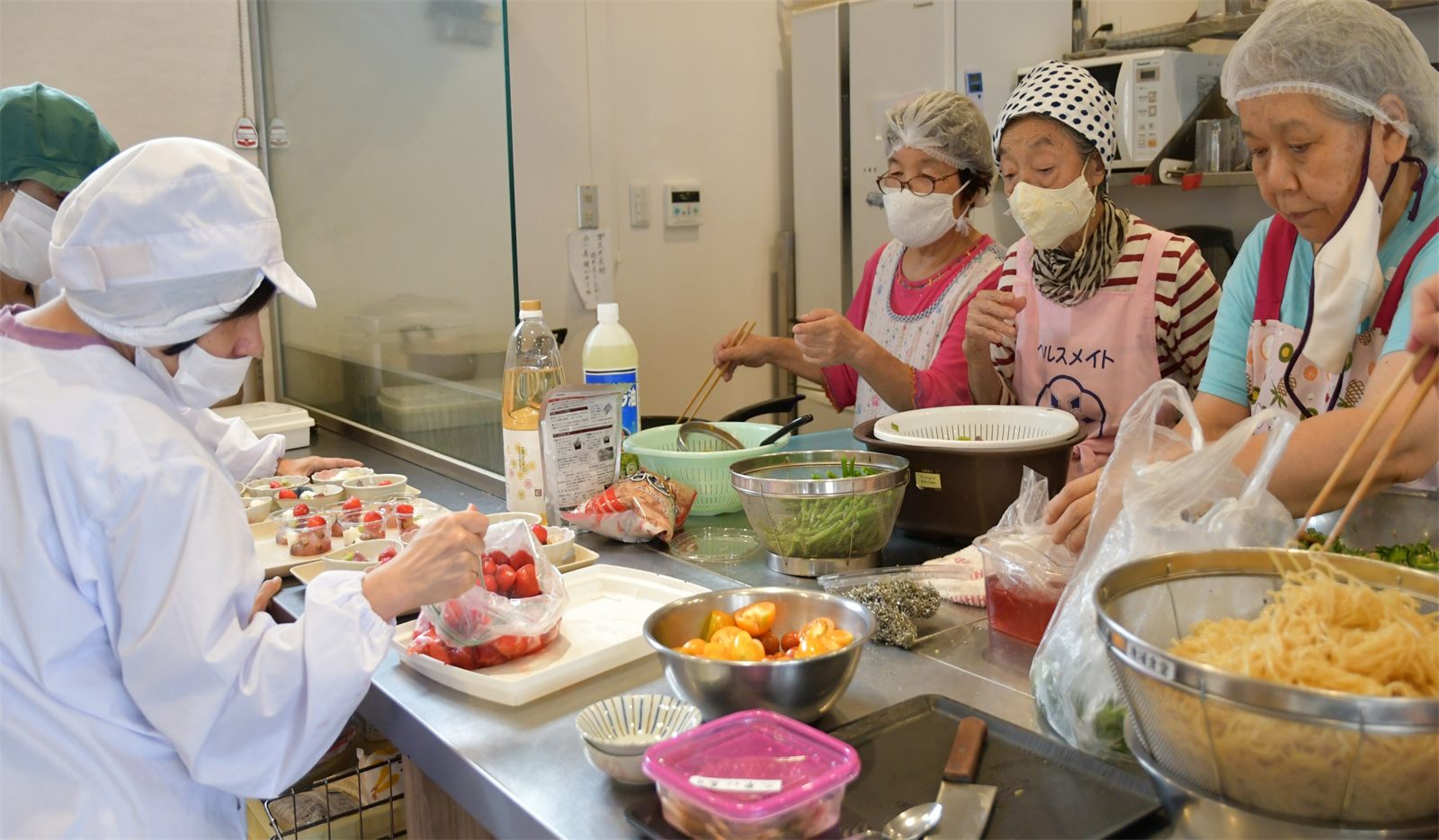 厨房でエプロン姿の方々が並んで、昼食のお弁当を仕込んでいる様子